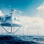 A hydrocarbon drilling platform rises above the surface of the sea. There are offshore wind turbines in the background.