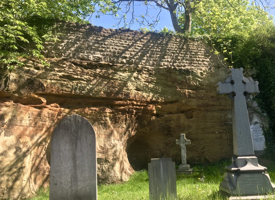 A yellow cliff with diagonal striations and a cave. A brick wall sit on top and there are gravestones in the grass at its base.