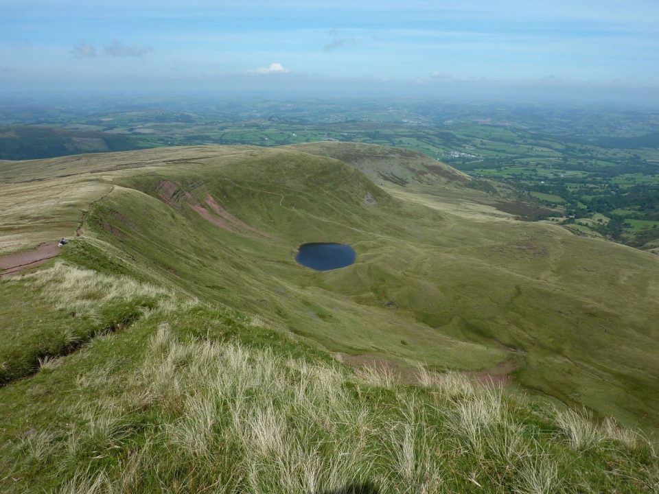 A curved, steep-sided, green grassy valley with an small, almost circular lake at the bottom of it. Flatter, lower land can be seen in the distance.