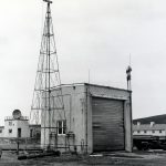 Black and white picture of a tall, triangular, metal framework next to a square building