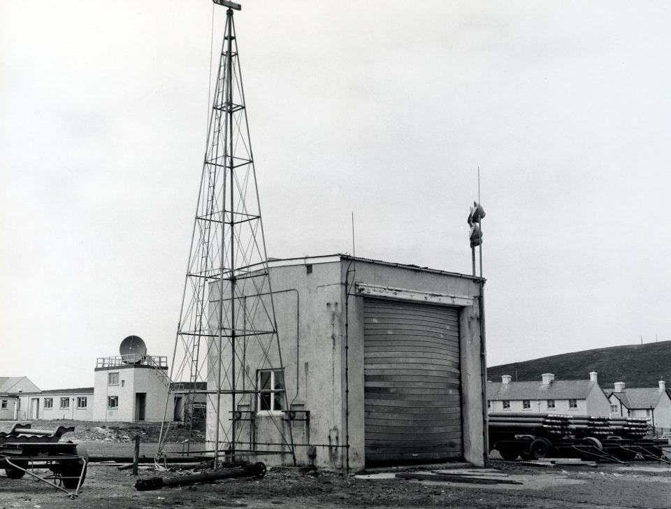 Black and white picture of a tall, triangular, metal framework next to a square building