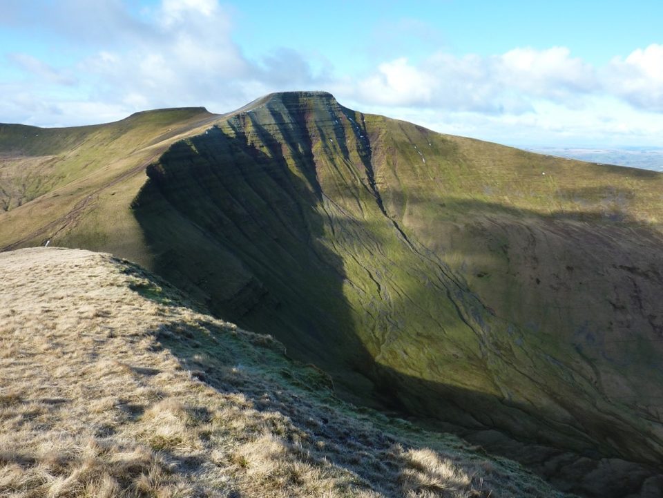 A curved, steep side of a hill showing interbedded, horizontal rocks. The top of the hill and another hill behind it are flat.