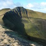 A curved, steep side of a hill showing interbedded, horizontal rocks. The top of the hill and another hill behind it are flat.