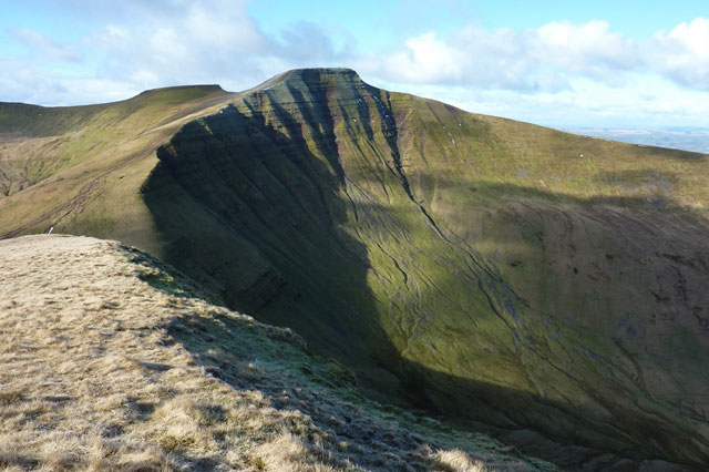 A curved, steep side of a hill showing interbedded, horizontal rocks. The top of the hill and another hill behind it are flat.