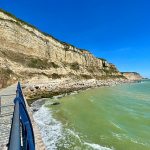 Cliffs with prominent horizontal beds stretch off into the distance above a rocky beach and the sea. There are blue railings and a cobbled walkway in the foregroud.