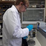 A white man in a white lab coat, safety glasses and blue rubber gloves is putting a substance into a glass jar