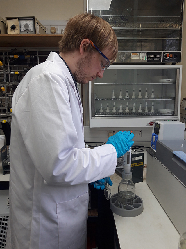 A white man in a white lab coat, safety glasses and blue rubber gloves is putting a substance into a glass jar