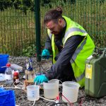 A bearded man in dark clothing, a hi-vis vest and blue latex gloves kneels on the ground in front of a fence. He is surrounded by sample pots containing liquids, an oil can, bottles, electrical wires and plastic boxes. He is dipping a sensor into one pot of liquid and looking at an electronic device in his hand.