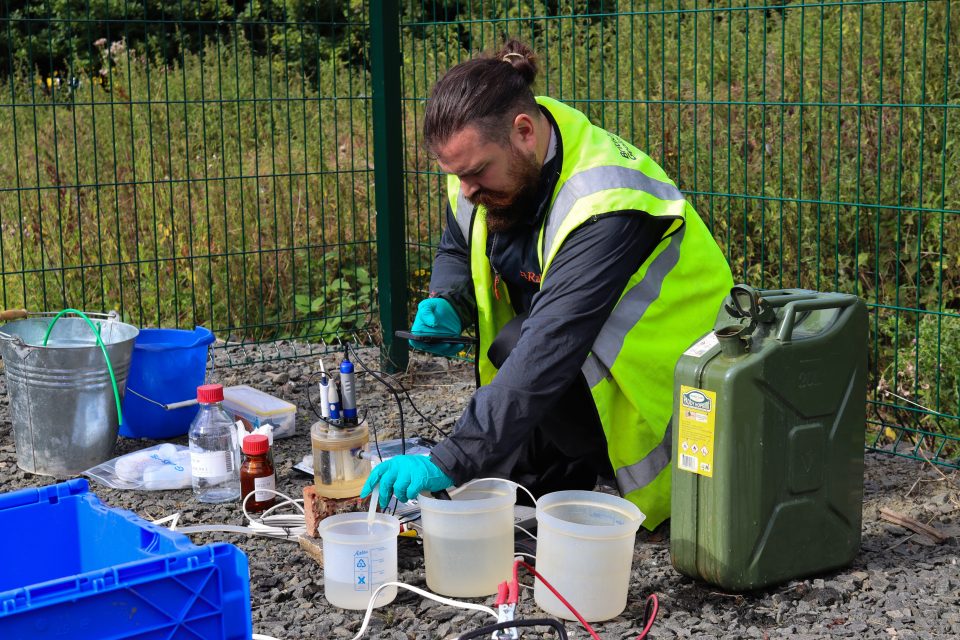 A bearded man in dark clothing, a hi-vis vest and blue latex gloves kneels on the ground in front of a fence. He is surrounded by sample pots containing liquids, an oil can, bottles, electrical wires and plastic boxes. He is dipping a sensor into one pot of liquid and looking at an electronic device in his hand.