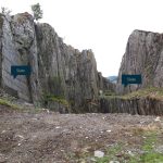 A quarry in grey rocks with steep sides of vertivslly layered rock on the left and in the centre, with a more rounded outcrop of rock on the right hand side