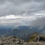 A view from the top of a mountain showing lower peaks and fistant lakes. The clouds are mostly below the altitude of the photo.
