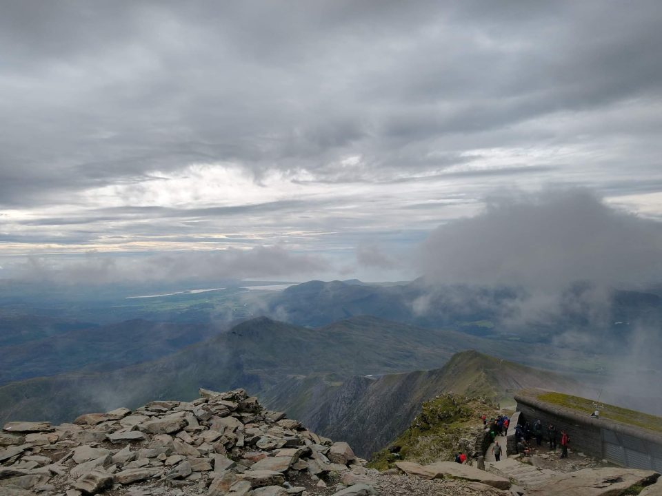 A view from the top of a mountain showing lower peaks and fistant lakes. The clouds are mostly below the altitude of the photo.