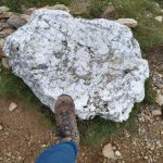 A white block of sock lying on bare brown soil. A foot wearing a brown boot is resting on the rock for scale. The foot is attached to a human who is wearing blue jeans.