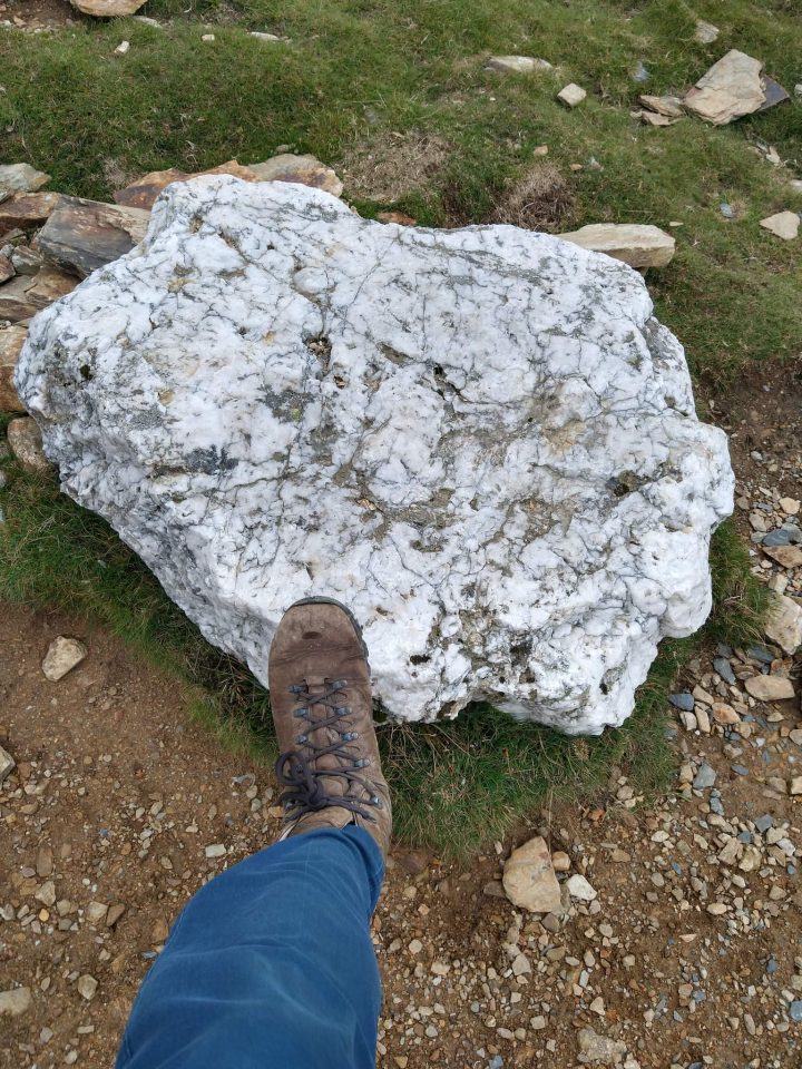 A white block of sock lying on bare brown soil. A foot wearing a brown boot is resting on the rock for scale. The foot is attached to a human who is wearing blue jeans.