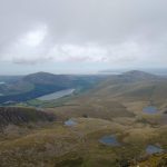 A view over peaks and lakes from the top of a high high. The sky is grey and cloudy.