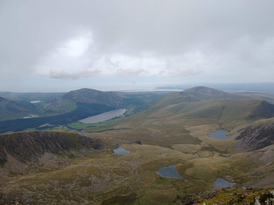 A view over peaks and lakes from the top of a high high. The sky is grey and cloudy.
