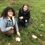 Two women crouch on grass in behind a couple of mushrooms. The woman on the left is pointing to the mushrooms and smiling.