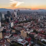 Drone image of Mexico City during sunset. There are many skyscrapers and blocks of smaller dwellings, many of which are a deep red or yellow in colour. Mountains can be seen in the background and some clouds dot the sky. David Arenas @ Pexels.
