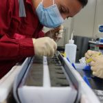 A woman wearing a medical face mask and a red jumpsuit bends over a long, semi-circular sediment sample. She is using long, thin tools to extract a bit of the sediment.