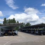 Cars parked in a car park beneath open-sided structures (car ports) with roofs covered in solar panels. The sky is bright, with big fluffy clouds.