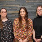 Three white women standing in front of a grey slate wall. Nicola on the left is wearing a black top with white flowers; she has long brown hair and glasses. Leah in the middle has long dark hair and is wearing an orange floral top. Angela on the right has bobbed blonde hair and is wearing glasses and a black jumper.