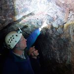 A geologist wearing blue overalls and a white hard hat points at the rock ceiling in a cave