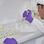 A white man wearing white PPE, glasses and purple nitrile gloves uses a long pipette to transfer liquid into a small bottle