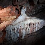 White crystalline rocks form speleothems that seem to flow over and drip from underlying pinkish rocks inside a cave.