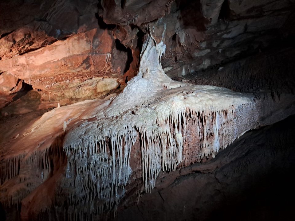 White crystalline rocks form speleothems that seem to flow over and drip from underlying pinkish rocks inside a cave.