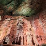 A wall of flowstone and speleothems 'dripping' down a cave wall. There a vertical stripes of pink, white and orange discolouration.