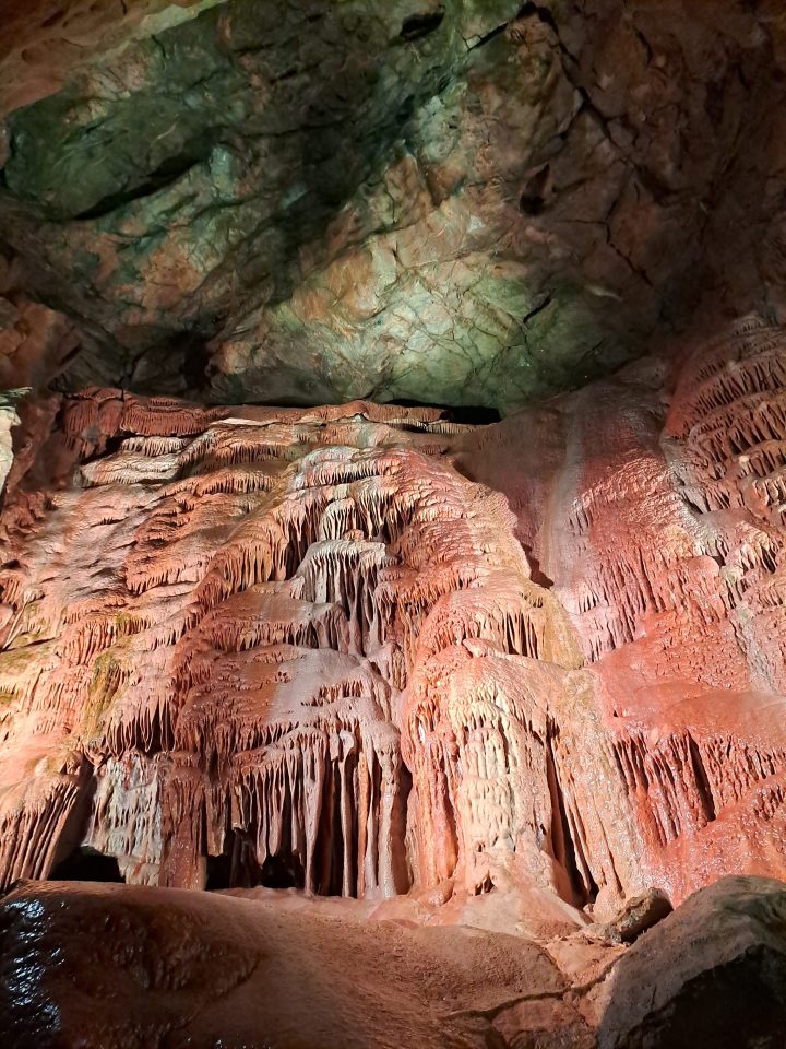 A wall of flowstone and speleothems 'dripping' down a cave wall. There a vertical stripes of pink, white and orange discolouration.