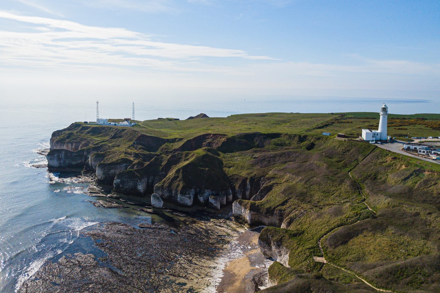 Cliffs of white chalk rock topped with green grass jut out into the sea. There are radio masts at the edge of the cliff and a lighthouse slightly inland. You can see rocks on the sea bed offshore from the beach at the bottom of the cliffs.