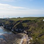 Cliffs of white chalk rock topped with green grass jut out into the sea. There are radio masts at the edge of the cliff and a lighthouse slightly inland. You can see rocks on the sea bed offshore from the beach at the bottom of the cliffs.