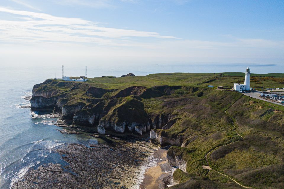 Cliffs of white chalk rock topped with green grass jut out into the sea. There are radio masts at the edge of the cliff and a lighthouse slightly inland. You can see rocks on the sea bed offshore from the beach at the bottom of the cliffs.