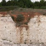 A white cliff overlain by reddish brown soil and green grass. There is a dip in the whiite rock that has been infilled by the red soil.