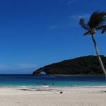 A tropical beach with white sand and two palm trees on the right. People are sunbathing and swimming in the sea. There is a distant, lushly vegetated hill with a rock arch in the distance.