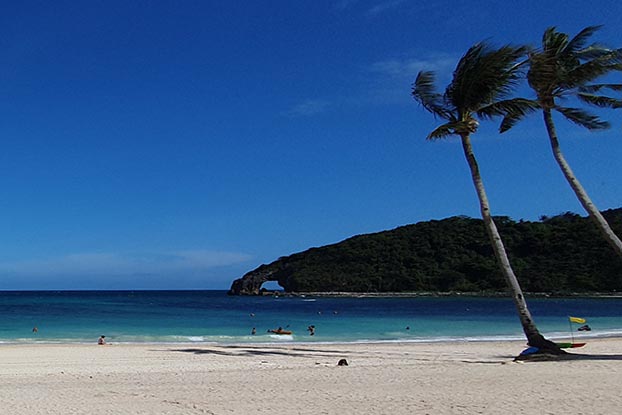 A tropical beach with white sand and two palm trees on the right. People are sunbathing and swimming in the sea. There is a distant, lushly vegetated hill with a rock arch in the distance.