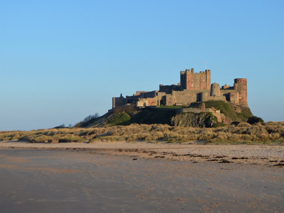 A castle sits on top of a high outcrop of rock. There and sand dunes and a beach in front of it.