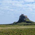 A castle sits on top of a high outcrop of rock. The rock rises from a flat, grassy plain.