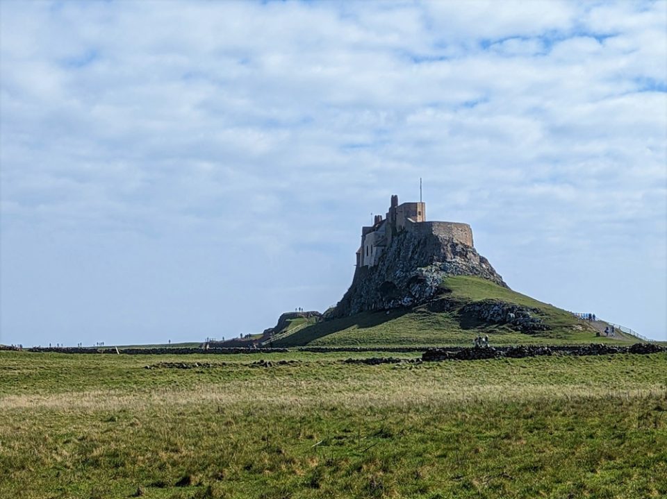 A castle sits on top of a high outcrop of rock. The rock rises from a flat, grassy plain.