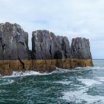 Three dark grey sea stack rising out of the sea. A brownish yellow band runs across the bottom of them.