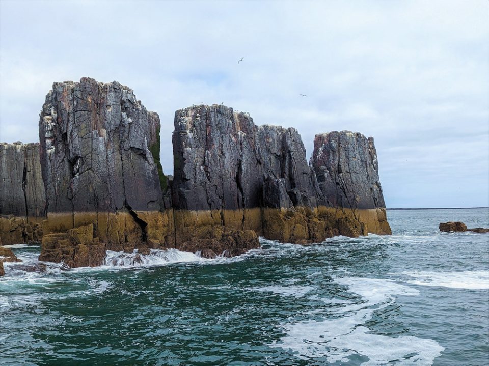 Three dark grey sea stack rising out of the sea. A brownish yellow band runs across the bottom of them.