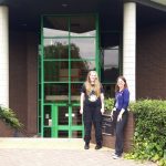 Two white women standing in front of a brown brick building with green window and door frames. One has long blonde hair and is wearing a black t shirt and trousers; the other has long brown hair and is wearing a blue top and black trousers. They are both smiling.