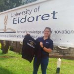 A woman stands in front of a sign for the University of Eldoret holding up a t shirt that says University of Waikato