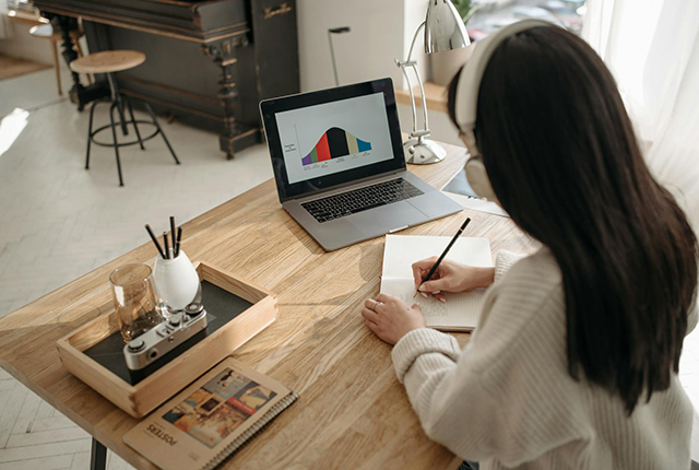 A young woman sits at a table with a laptop on it and a tray with pens and other stationery. She is writing on a pad of paper.