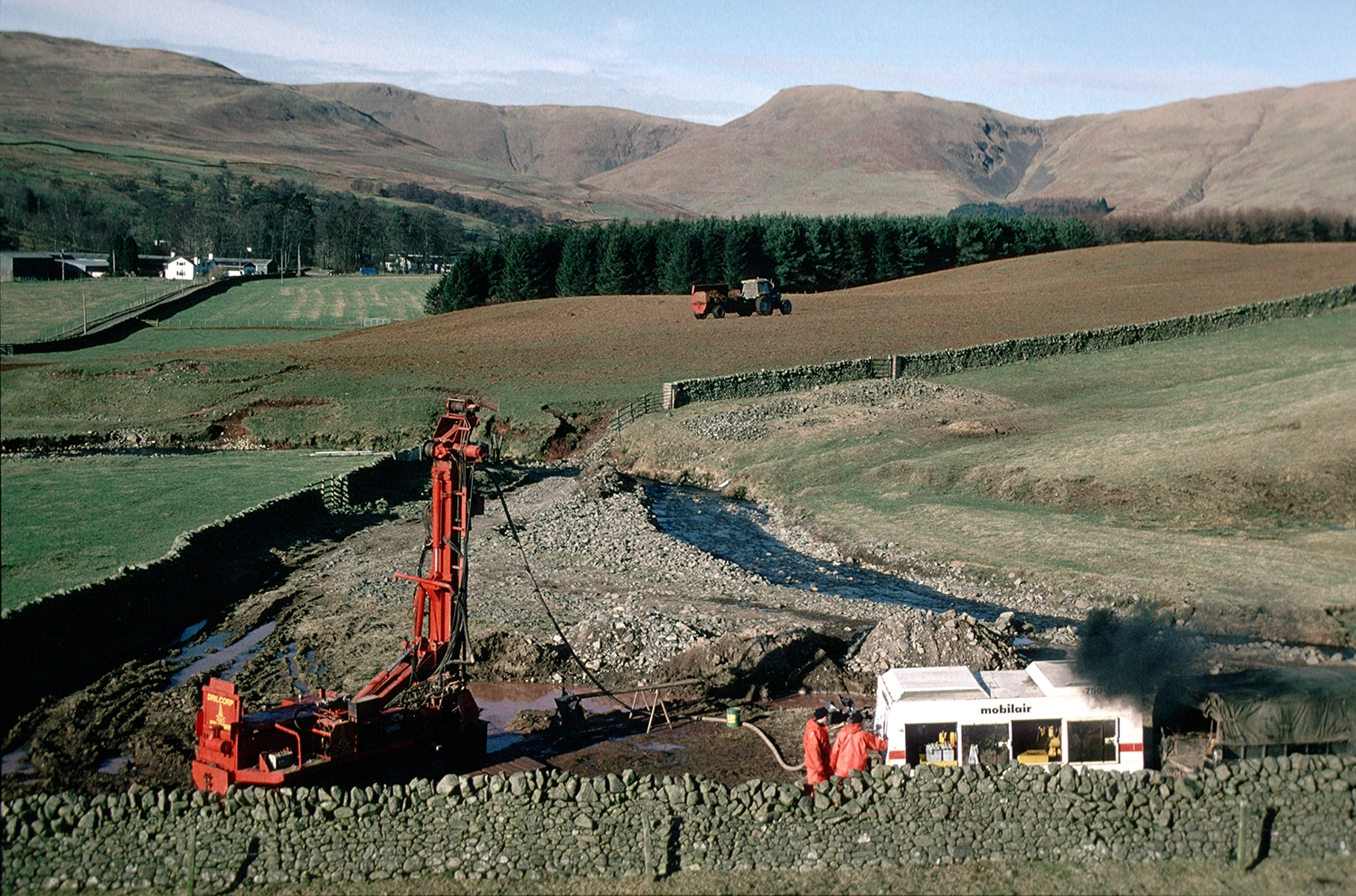 A borehole being drilled to assess groundwater aquifer potential in Dumfries and Galloway, Scotland. Records state that 5 metres of coarse superficial gravel was present atop Permian bedrock at this site. The drill site lies in the Till Dominant Domain, whilst the Mountain and Valley Domain is visible in the background of the image.