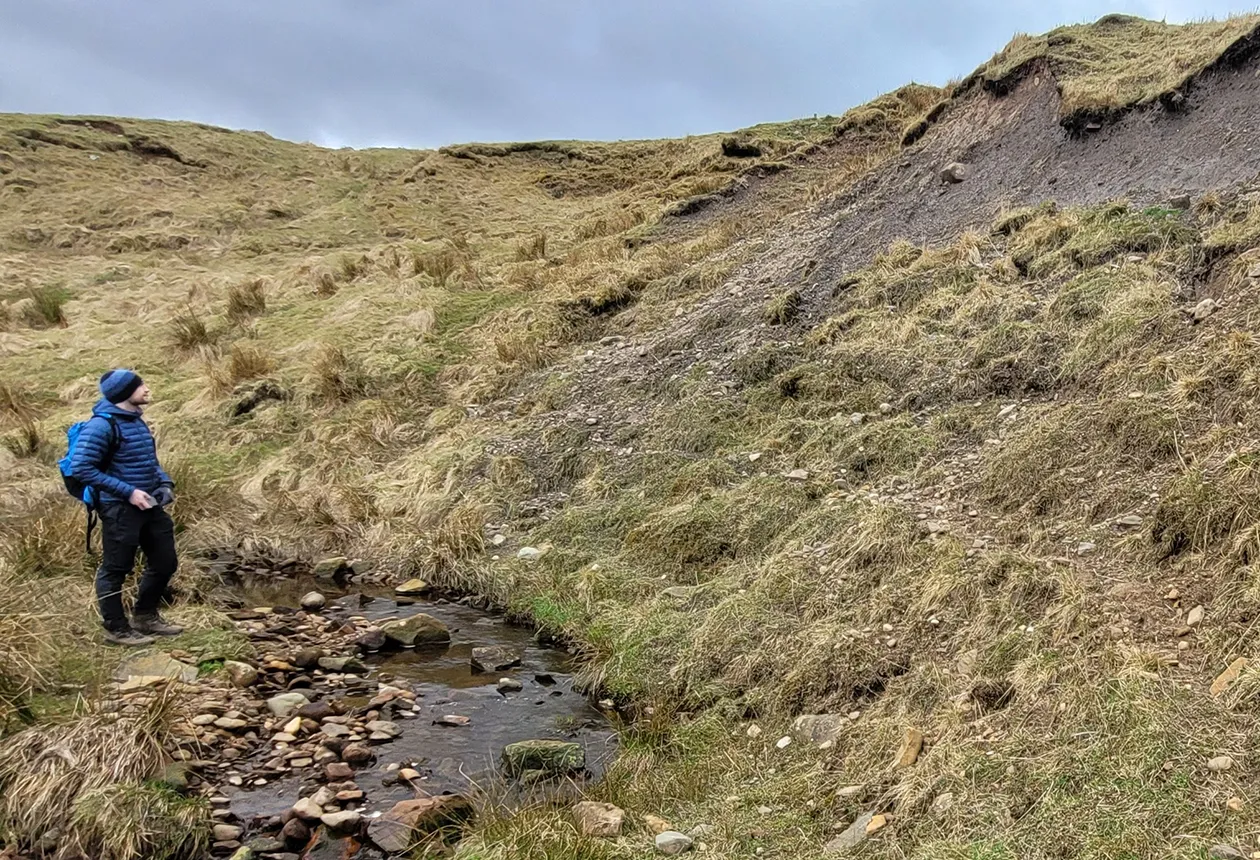 BGS Marine Geoscientist Duncan Stevens observing the glacial till exposure at the Black Burn site, Auchencorth Moss. The site shows multiple stacked till units with varying composition and structure, offering valuable insights into sediment heterogeneity, a key consideration when interpreting offshore stratigraphy and designing marine infrastructure. BGS © UKRI.