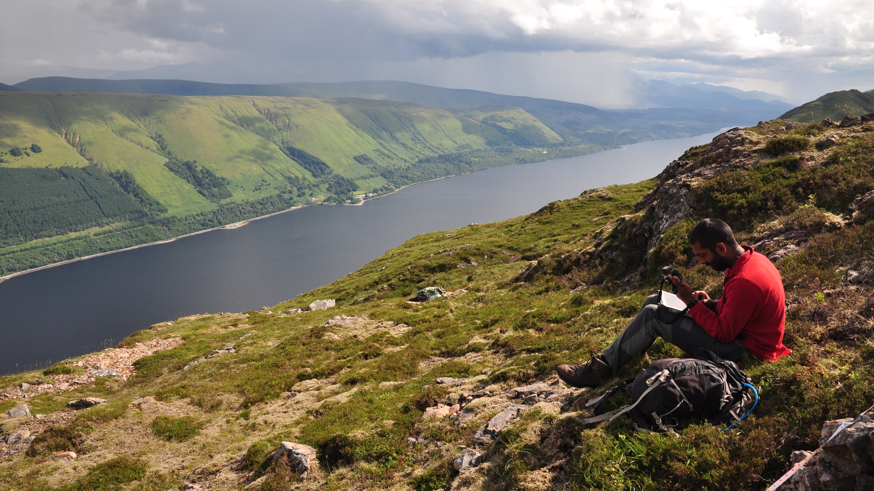 A view over a lake from a hilllside. A man wearing a red top and grey trousers is sitting on the ground writing something in a notebook. There is a rucksack next to him.