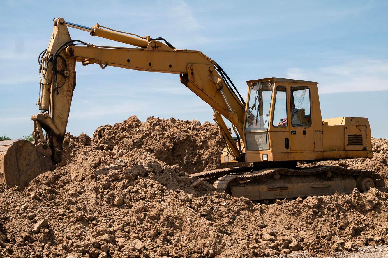 A large yellow digger excavating brown soil on top of a soil heap.