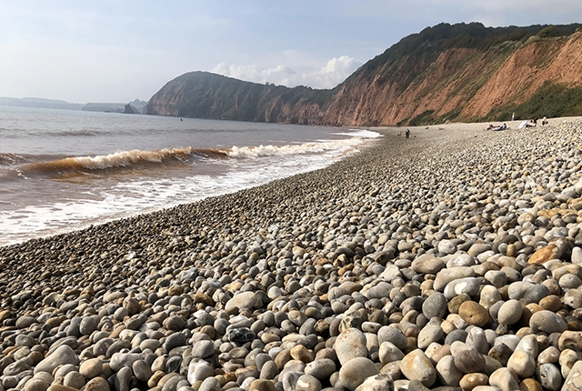 View of a pebbly beach, with cliffs in teh background and waves breaking on the shore. Some people are walking in the shoreline in the distance, others are sitting on the pebbles. BGS © UKRI